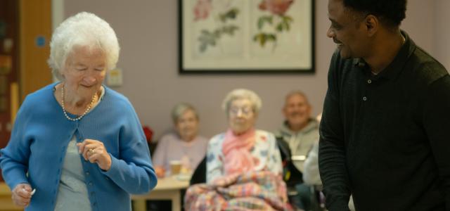 Elderly woman dances with caregiver in a community room by Age Cymru courtesy of Unsplash.